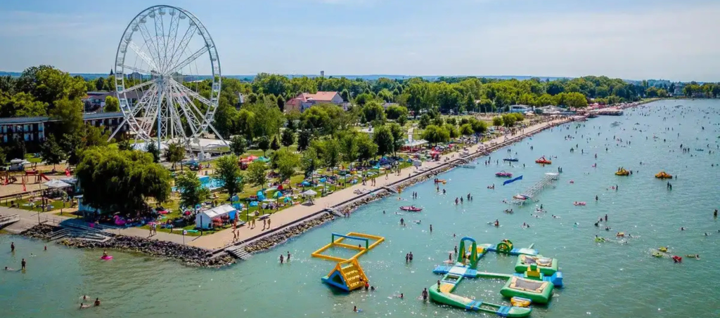 Summer day at Siófok Beach on Lake Balaton, Hungary, with the Siófok Eye Ferris wheel, water park inflatables, and crowds enjoying swimming and sunbathing.