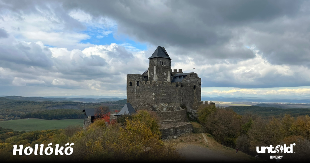 The ancient Hollókő Castle standing proudly on a hilltop, framed by golden autumn trees and a cloudy blue sky.