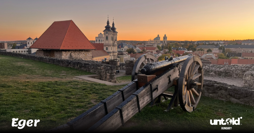 The medieval Eger Castle at sunset with warm golden light, red rooftops, and a cannon viewpoint overlooking the old town.